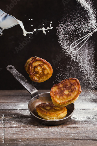 Pancakes and frying pan levitate over a table, and on top a whisk and sprinkled with flour. Milk is pouring from a cup