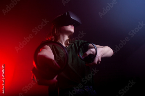 girl gamer in modern virtual reality glasses plays a shooter on a dark red-blue background, a woman shoots with game joysticks © Богдан Маліцький