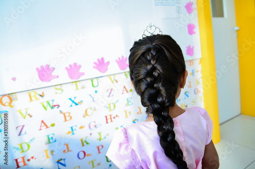 girl on her back with braid in her hair and pink dress,child with hair braid, child learning alphabet at classroom board, girl seen from behind in early education, student interacting with letters 