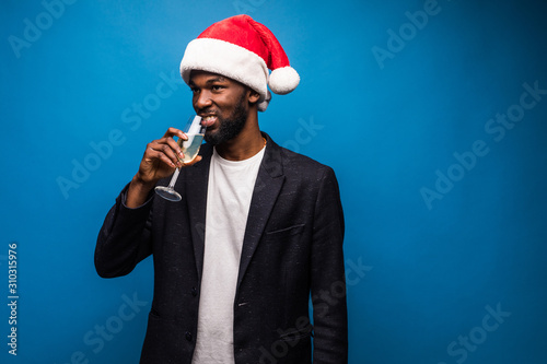 Christmas time noel eve festive. Handsome african cman in headwear formal wear suit jacket in santa hat drink champagne isolated on blue background © F8  \ Suport Ukraine