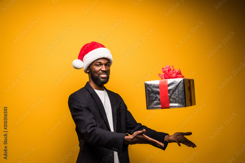 African American man in Santa hat throwing up gift box isolated on yellow background African American man in Santa hat throwing up gift box isolated on yellow background