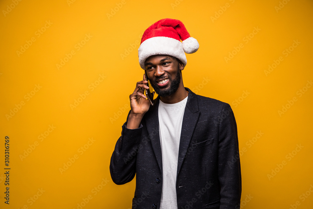 Young african man in santa hat talking on the phone isolated on yellow background Young african man in santa hat talking on the phone isolated on yellow background
