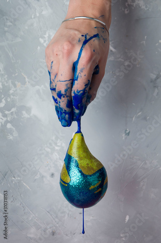 The female hand holds a ripe yellow pear in blue paint with spangles. A drop drains from the fruit. On a textured gray background.