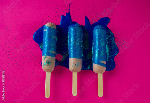 Sausages on sticks in blue sparkle paint. On a pink background, top view photo.