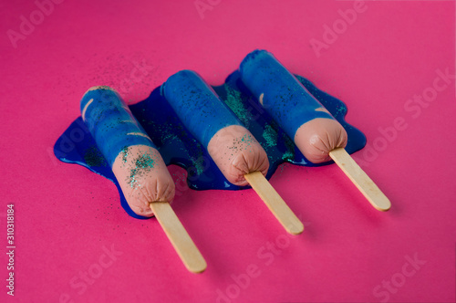 Sausages on sticks in blue sparkle paint. On a pink background, top view photo.