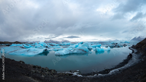 Panoramic of Jokulsarlon glacier lagoon covered with blue icebergs, dramatic landscape, some people walk near the shore