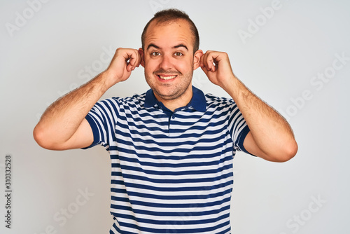 Young man wearing casual striped polo standing over isolated white background Smiling pulling ears with fingers, funny gesture. Audition problem