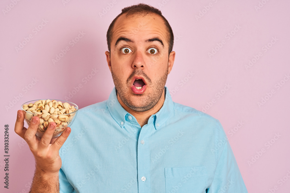 Young man holding bowl with pistachios standing over isolated pink background scared in shock with a surprise face, afraid and excited with fear expression Young man holding bowl with pistachios standing over isolated pink background scared in shock with a surprise face, afraid and excited with fear expression