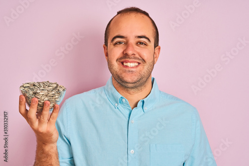 Young man holding bowl with sunflowers seeds standing over isolated pink background with a happy face standing and smiling with a confident smile showing teeth