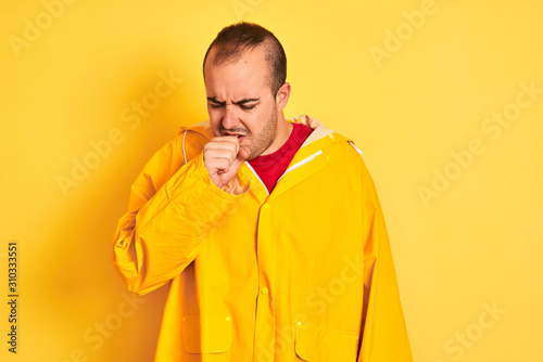 Young man wearing rain coat standing over isolated yellow background feeling unwell and coughing as symptom for cold or bronchitis. Healthcare concept.