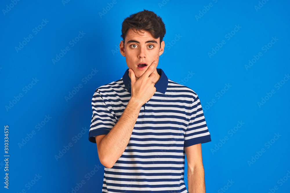 Teenager boy wearing casual t-shirt standing over blue isolated background Looking fascinated with disbelief, surprise and amazed expression with hands on chin Teenager boy wearing casual t-shirt standing over blue isolated background Looking fascinated with disbelief, surprise and amazed expression with hands on chin