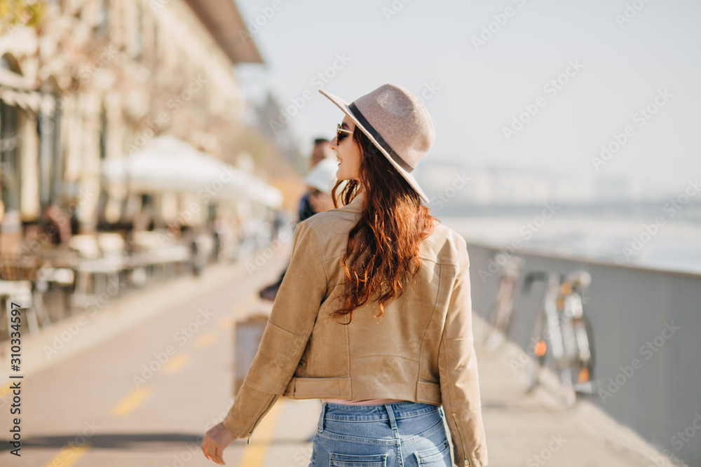 Portrait from back of stylish slim woman with long red hair walking down the street. Graceful ginger girl in beige jacket looking around while exploring town.