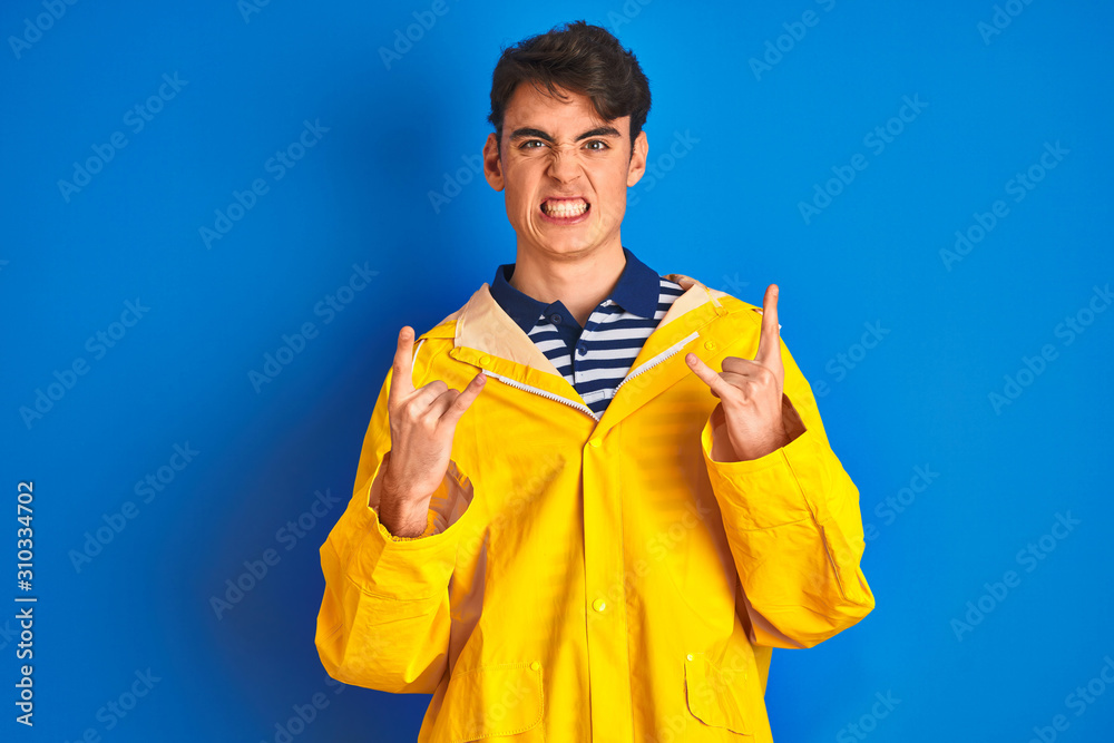 Teenager fisherman boy wearing yellow raincoat over isolated background shouting with crazy expression doing rock symbol with hands up. Music star. Heavy concept. Teenager fisherman boy wearing yellow raincoat over isolated background shouting with crazy expression doing rock symbol with hands up. Music star. Heavy concept.