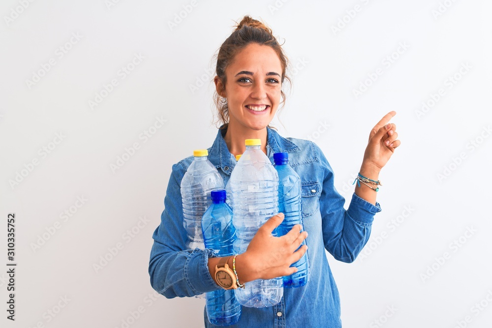 Young redhead woman holding plastic bottle to recycle over isolated background very happy pointing with hand and finger to the side Young redhead woman holding plastic bottle to recycle over isolated background very happy pointing with hand and finger to the side