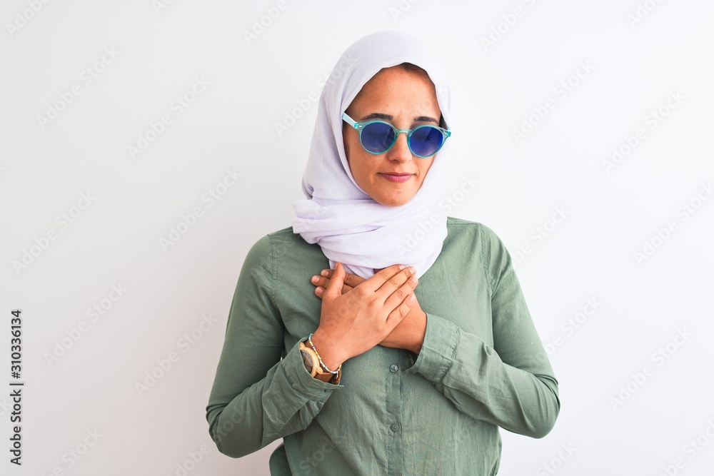 Young Arab woman wearing hijab and summer sunglasses over isolated background smiling with hands on chest with closed eyes and grateful gesture on face. Health concept.