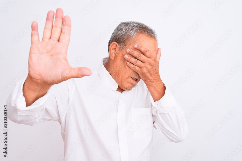 Senior grey-haired man wearing elegant shirt standing over isolated white background covering eyes with hands and doing stop gesture with sad and fear expression. Embarrassed and negative concept. Senior grey-haired man wearing elegant shirt standing over isolated white background covering eyes with hands and doing stop gesture with sad and fear expression. Embarrassed and negative concept.