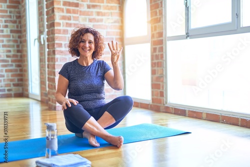 Middle age beautiful sportswoman wearing sportswear sitting on mat practicing yoga at home showing and pointing up with fingers number four while smiling confident and happy.