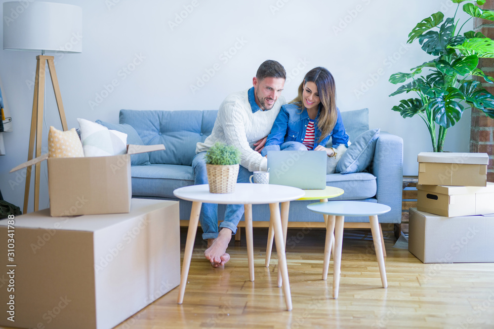 Young beautiful couple sitting on the sofa drinking cup of coffee using laptop at new home around cardboard boxes Young beautiful couple sitting on the sofa drinking cup of coffee using laptop at new home around cardboard boxes