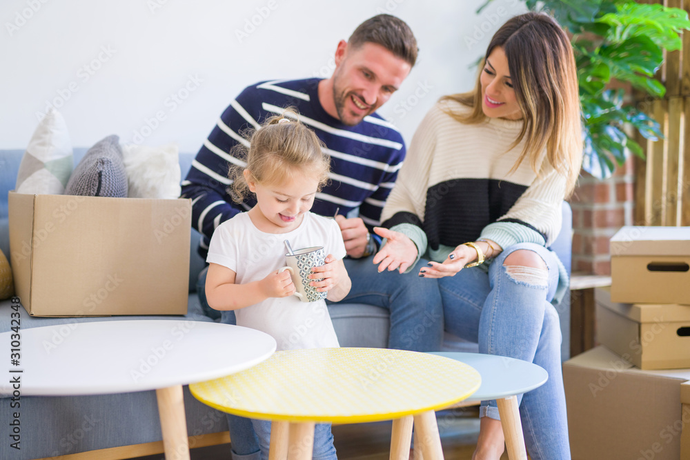 Beautiful family, parents sitting on the sofa drinking coffee looking his kid playing at new home around cardboard boxes