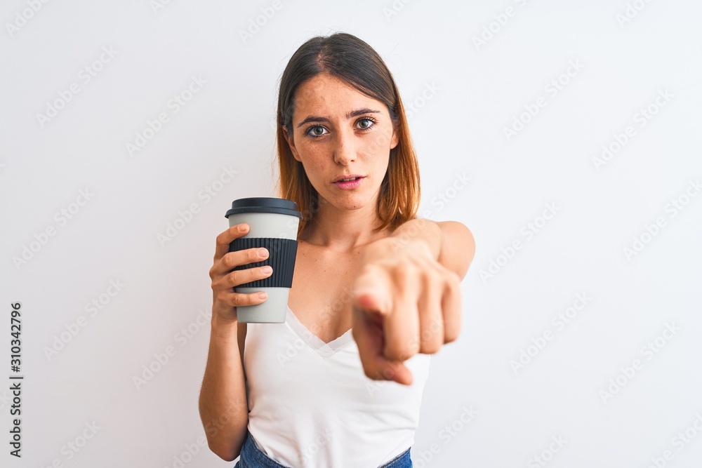 Beautiful redhead woman drinking take away coffee over isolated background pointing with finger to the camera and to you, hand sign, positive and confident gesture from the front Beautiful redhead woman drinking take away coffee over isolated background pointing with finger to the camera and to you, hand sign, positive and confident gesture from the front