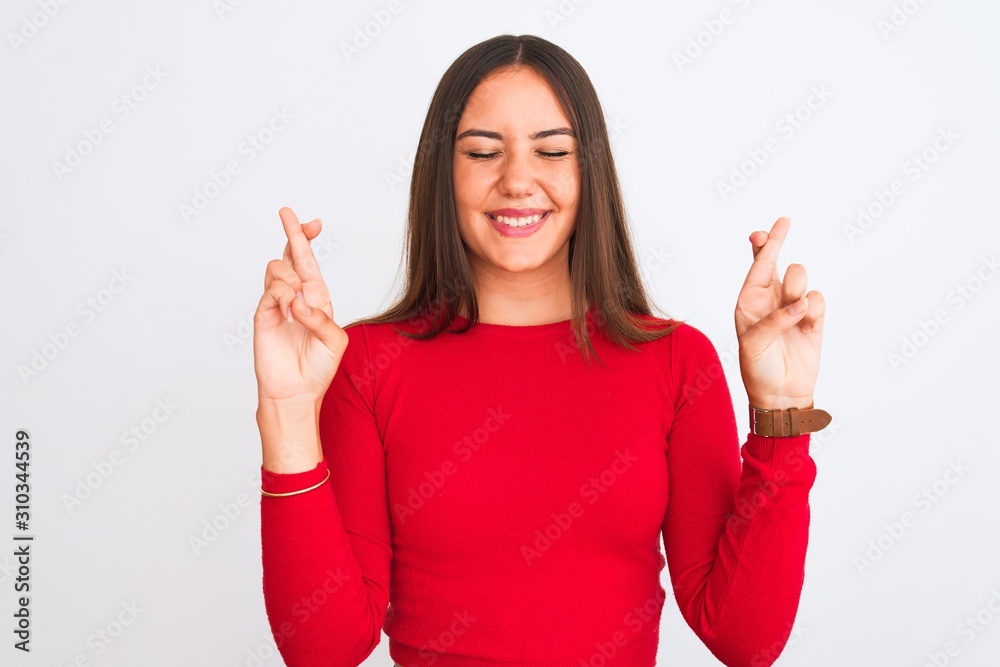 Young beautiful girl wearing red casual t-shirt standing over isolated white background gesturing finger crossed smiling with hope and eyes closed. Luck and superstitious concept. Young beautiful girl wearing red casual t-shirt standing over isolated white background gesturing finger crossed smiling with hope and eyes closed. Luck and superstitious concept.