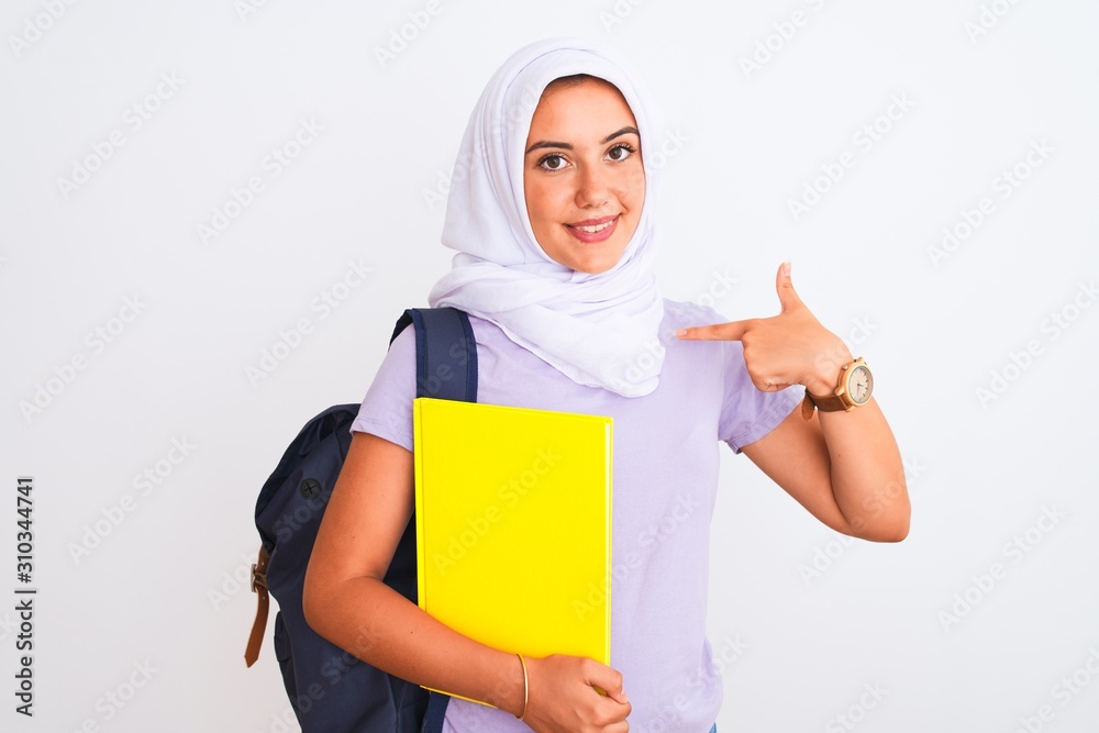 Arab student girl wearing hijab and backpack holding book over isolated white background with surprise face pointing finger to himself Arab student girl wearing hijab and backpack holding book over isolated white background with surprise face pointing finger to himself