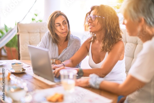 Meeting of middle age women having lunch and drinking coffee. Mature friends smiling happy using laptop at home on a sunny day