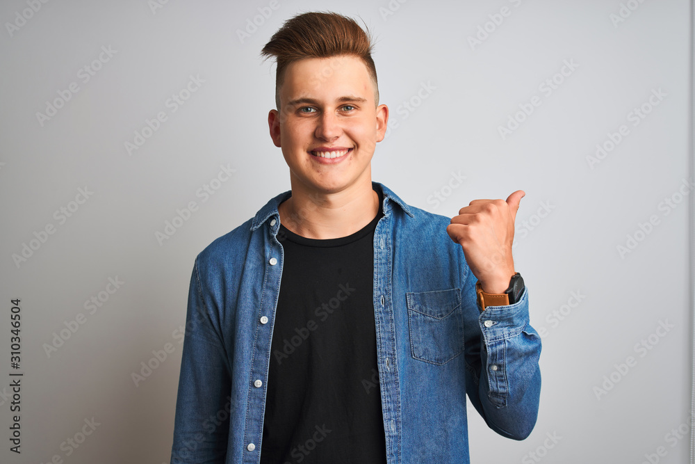 Young handsome man wearing denim shirt standing over isolated white background smiling with happy face looking and pointing to the side with thumb up. Young handsome man wearing denim shirt standing over isolated white background smiling with happy face looking and pointing to the side with thumb up.