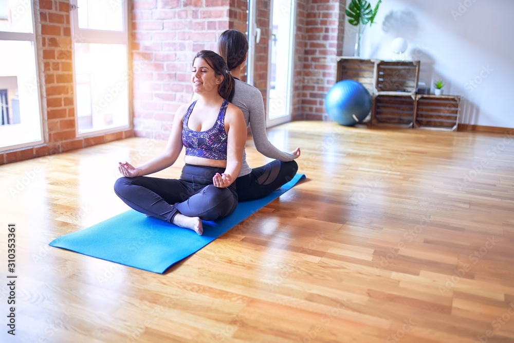 2 young beautiful sportswomen practicing yoga. Sitting on the floor doing lotus pose at gym