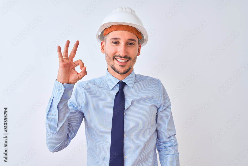Young business man wearing contractor safety helmet over isolated background smiling positive doing ok sign with hand and fingers. Successful expression.