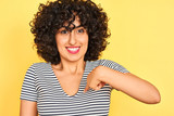Young arab woman with curly hair wearing striped dress over isolated yellow background with surprise face pointing finger to himself