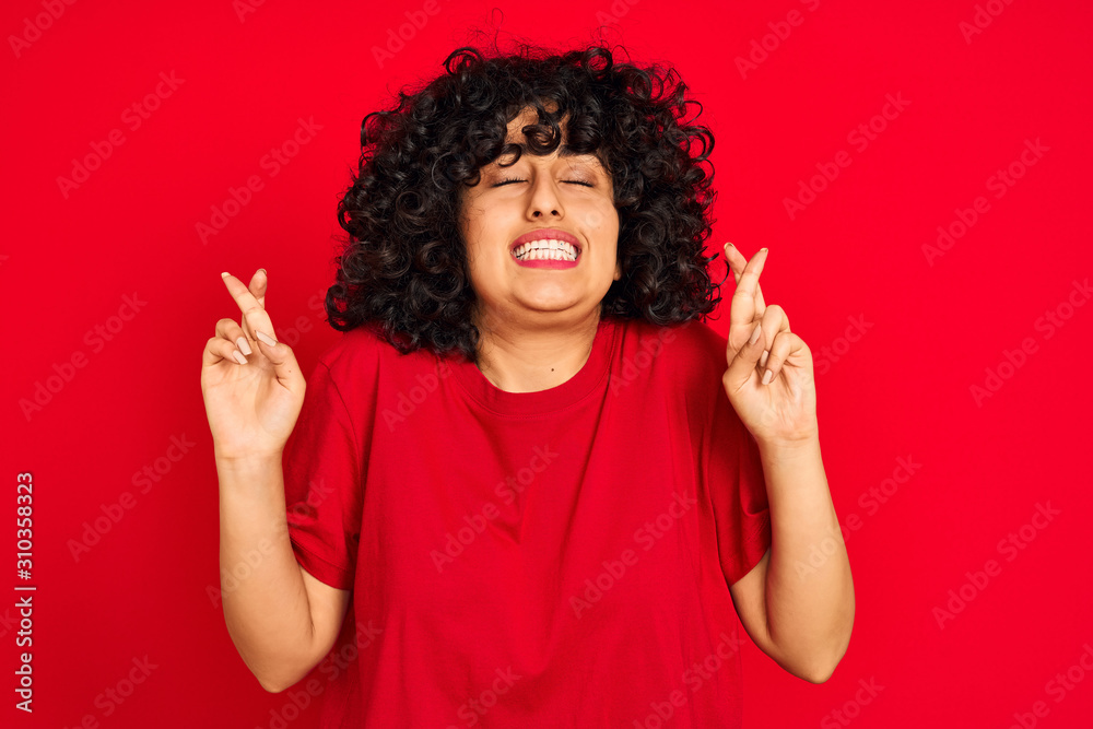 Young arab woman with curly hair wearing casual t-shirt over isolated red background gesturing finger crossed smiling with hope and eyes closed. Luck and superstitious concept. Young arab woman with curly hair wearing casual t-shirt over isolated red background gesturing finger crossed smiling with hope and eyes closed. Luck and superstitious concept.