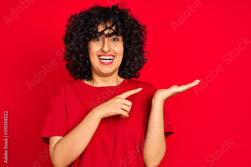 Young arab woman with curly hair wearing casual t-shirt over isolated red background amazed and smiling to the camera while presenting with hand and pointing with finger.