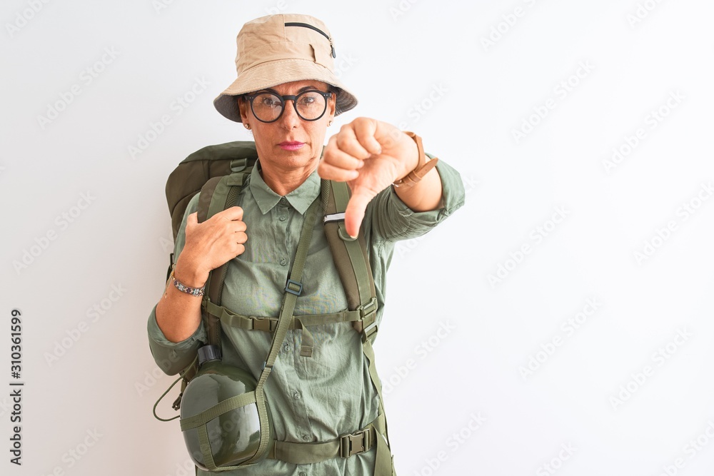 Middle age hiker woman wearing backpack canteen hat glasses over isolated white background looking unhappy and angry showing rejection and negative with thumbs down gesture. Bad expression. Middle age hiker woman wearing backpack canteen hat glasses over isolated white background looking unhappy and angry showing rejection and negative with thumbs down gesture. Bad expression.