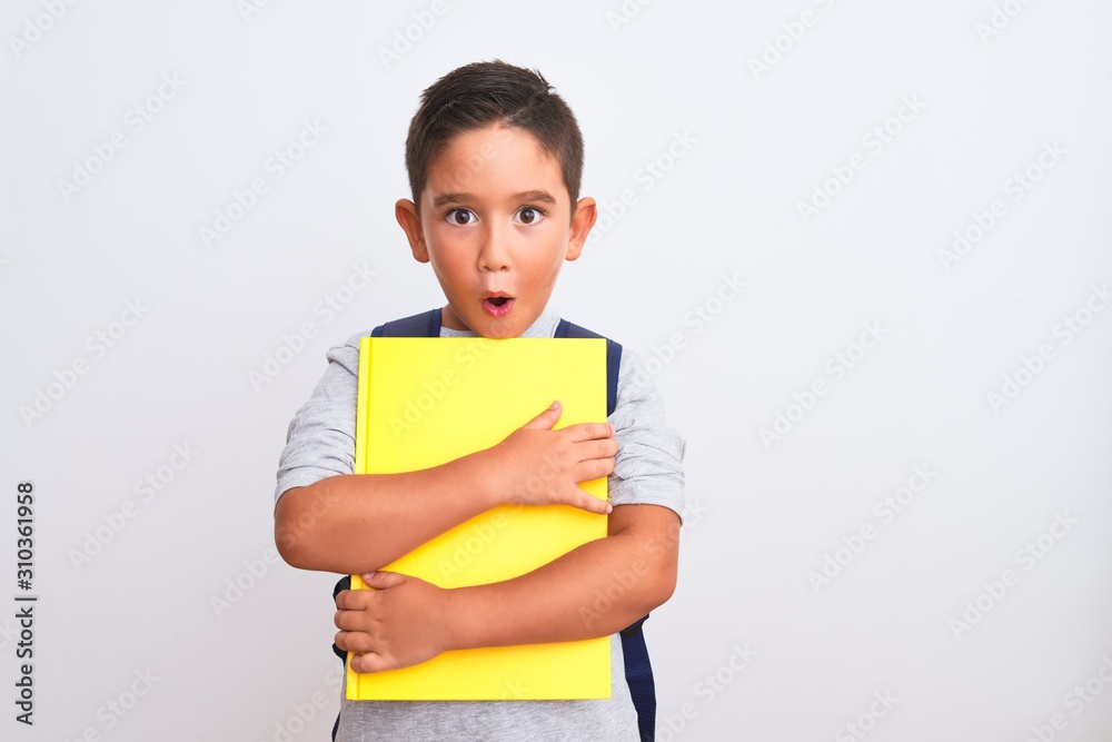 Beautiful student kid boy wearing backpack holding book over isolated white background scared in shock with a surprise face, afraid and excited with fear expression Beautiful student kid boy wearing backpack holding book over isolated white background scared in shock with a surprise face, afraid and excited with fear expression