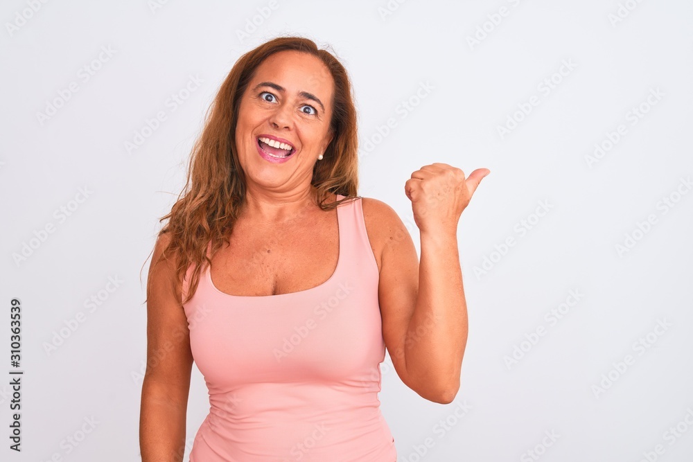 Middle age mature woman standing over white isolated background smiling with happy face looking and pointing to the side with thumb up.