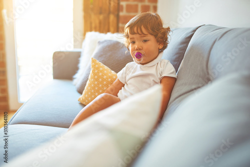 Beautiful toddler child girl wearing white t-shirt sitting on the sofa using pacifier