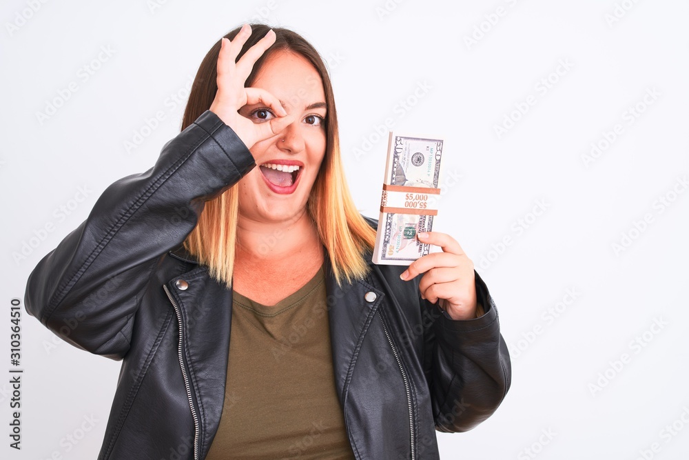 Young beautiful woman holding dollars standing over isolated white background with happy face smiling doing ok sign with hand on eye looking through fingers Young beautiful woman holding dollars standing over isolated white background with happy face smiling doing ok sign with hand on eye looking through fingers