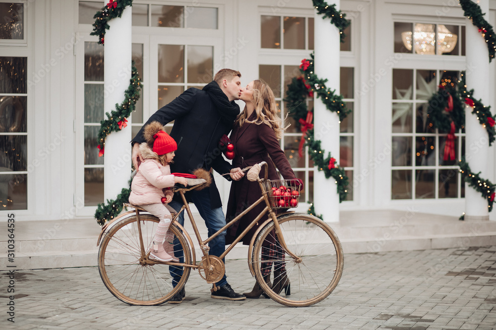 Obraz premium Stock photo of loving husband and wife kissing while holding a golden bicycle with their little daughter in red hait on it. They are kissing against beautifully decorated white house with Christmas