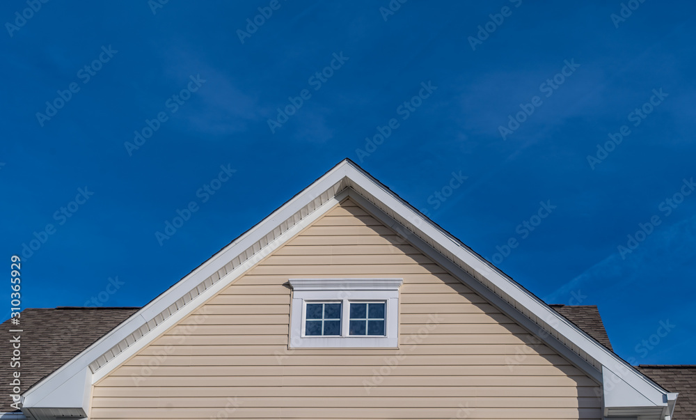Single gable close up with beige vinyl siding on luxury single family residential home with double attic window in white frame with blue sky background