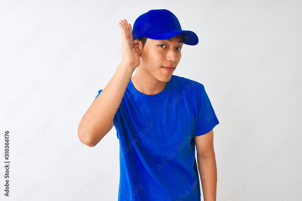 Chinese deliveryman wearing blue t-shirt and cap standing over isolated white background smiling with hand over ear listening an hearing to rumor or gossip. Deafness concept.