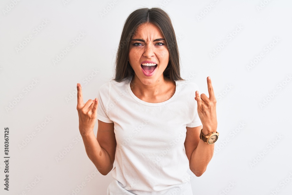 Young beautiful woman wearing casual t-shirt standing over isolated white background shouting with crazy expression doing rock symbol with hands up. Music star. Heavy music concept. Young beautiful woman wearing casual t-shirt standing over isolated white background shouting with crazy expression doing rock symbol with hands up. Music star. Heavy music concept.