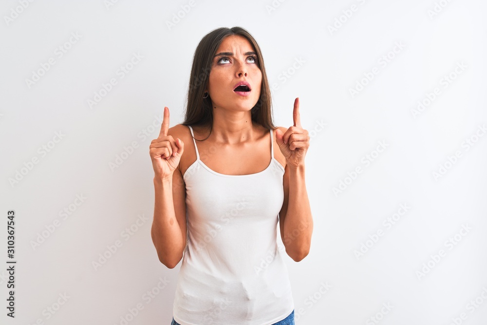 Young beautiful woman wearing casual t-shirt standing over isolated white background amazed and surprised looking up and pointing with fingers and raised arms. Young beautiful woman wearing casual t-shirt standing over isolated white background amazed and surprised looking up and pointing with fingers and raised arms.