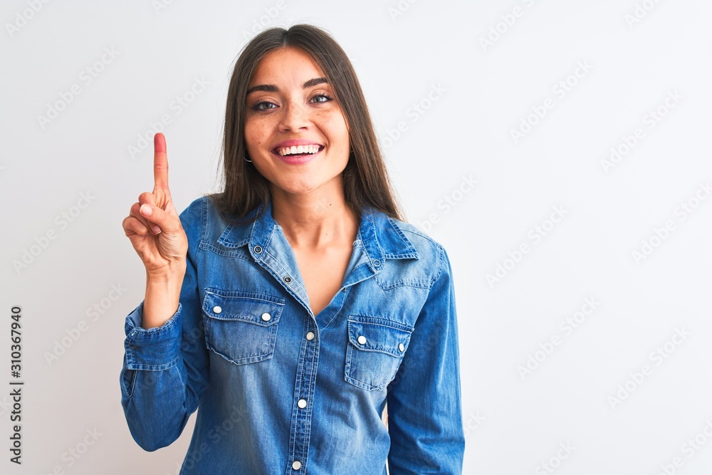 Young beautiful woman wearing casual denim shirt standing over isolated white background showing and pointing up with finger number one while smiling confident and happy. Young beautiful woman wearing casual denim shirt standing over isolated white background showing and pointing up with finger number one while smiling confident and happy.