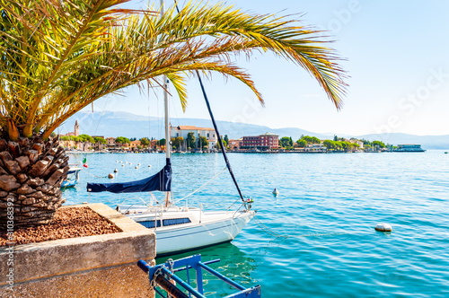 Garda lake western shore, Lombardy Italy. View on Toscolano Maderno cityscape through palm leaves growing on the beach with parked white sailing yacht on crystal clear blue water of the amazing lake