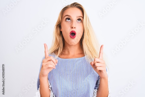 Young beautiful woman wearing elegant blue t-shirt standing over isolated white background amazed and surprised looking up and pointing with fingers and raised arms.