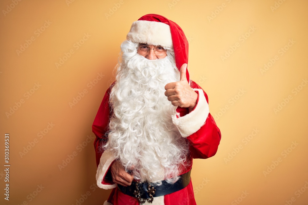 Middle age handsome man wearing Santa costume standing over isolated yellow background doing happy thumbs up gesture with hand. Approving expression looking at the camera with showing success.