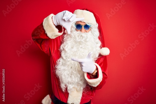 Middle age man wearing Santa Claus costume and sunglasses over isolated red background smiling making frame with hands and fingers with happy face. Creativity and photography concept.