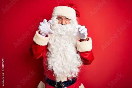 Middle age handsome man wearing Santa costume standing over isolated red background smiling funny doing claw gesture as cat, aggressive and sexy expression