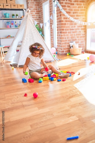 Beautiful toddler wearing glasses and unicorn diadem sitting playing with building blocks at kindergarten